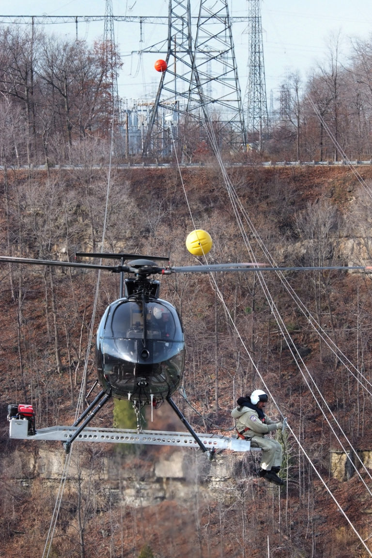 An individual works on electric wiring from the base of a helicopter with trees in the background.